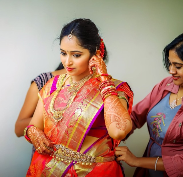 Bride in an orange silk saree being helped with her outfit